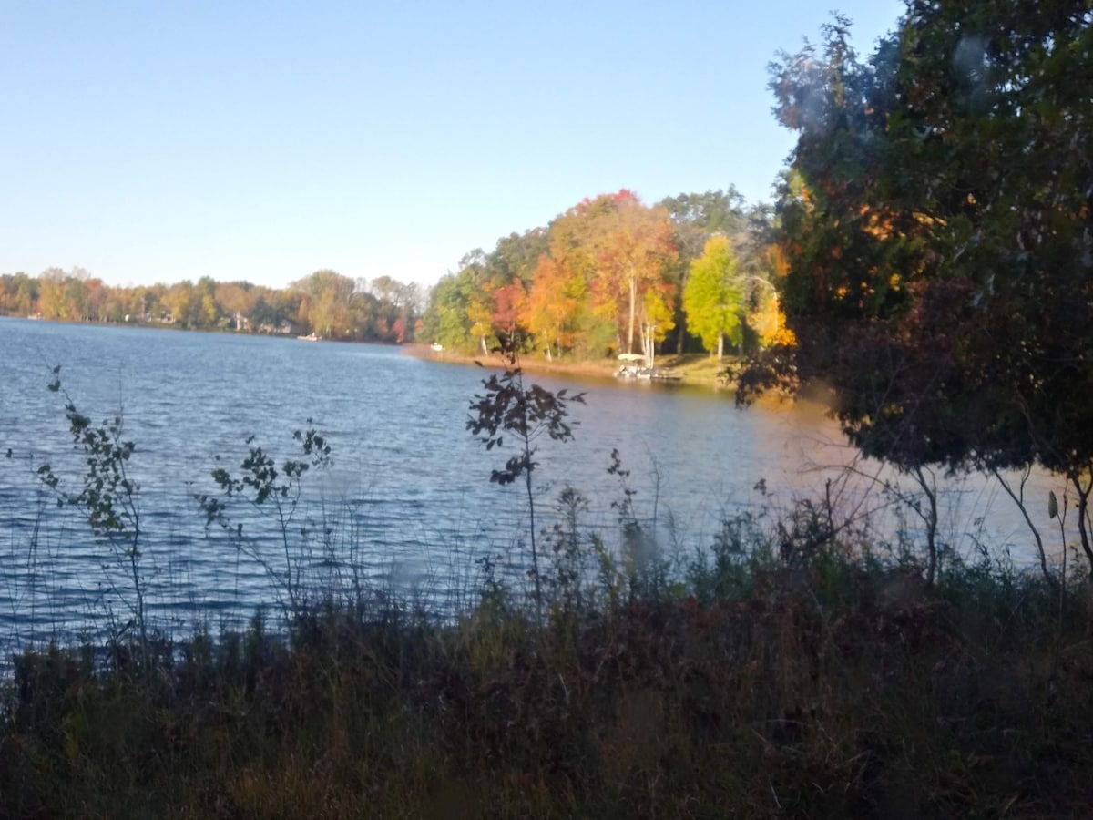 A serene view of Half Moon Lake captures the calm water reflecting surrounding trees in various autumnal colors. The shoreline features a mix of greenery and colorful foliage, providing a natural frame for the tranquil scene.