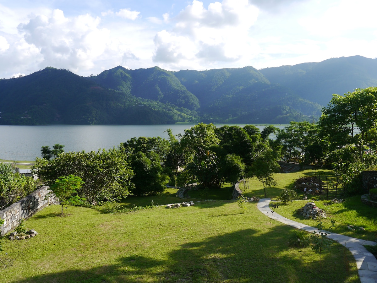 A verdant garden leads down to the serene Lake Phéwa, framed by lush green hills in the background. A winding stone path meanders through the grass, inviting exploration alongside the picturesque scene of water and mountains.