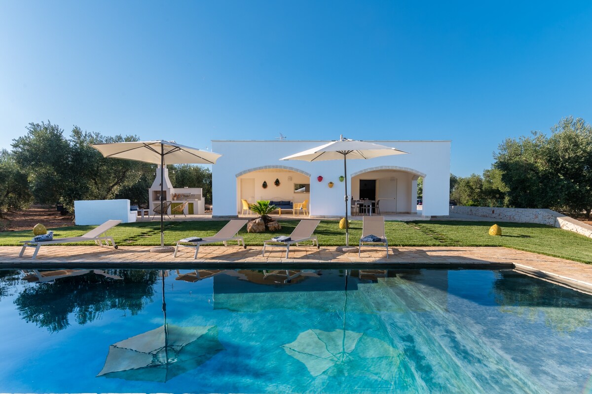 A private pool with clear water reflects the blue sky, surrounded by lounge chairs and sun umbrellas. A spacious outdoor area features a well-maintained lawn and backs onto a modern white building with arched windows. Olive trees create a serene backdrop.