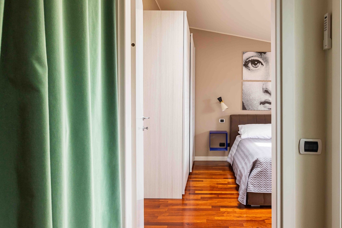 The bedroom is accessed through an entrance framed by curtains. Features include a spacious bed with a patterned cover, a double wardrobe, and a bedside table. A wall art piece of a face is displayed above the bed, complementing the natural wooden flooring.