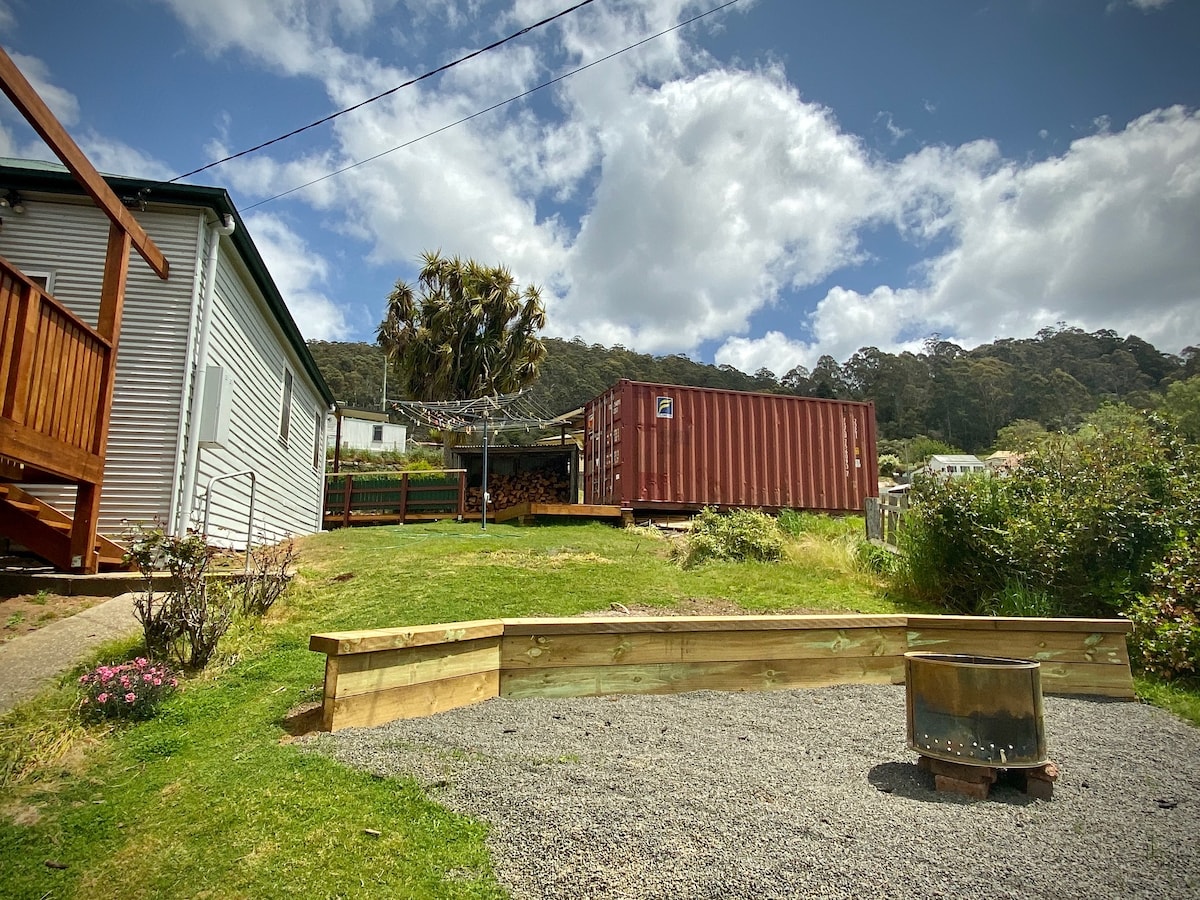 A spacious outdoor area features a well-maintained lawn and flower beds, surrounded by greenery. A wooden retaining wall borders the space. A fire pit is positioned on gravel, with a shipping container visible in the background under a partly cloudy sky.