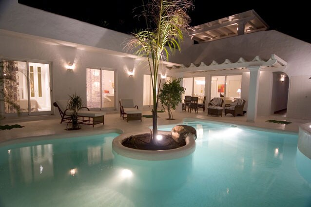 A well-lit outdoor area features a serene pool with gentle lighting, surrounded by palm trees and comfortable lounge chairs. Glass doors open into the interior, revealing hints of warm decor and cozy furniture in the background.