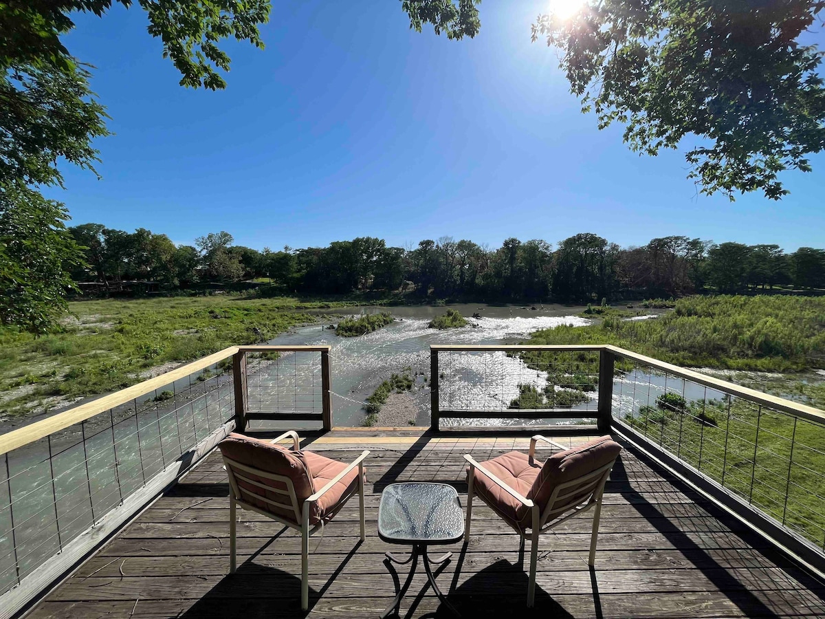A serene outdoor space is visible with two lawn chairs positioned around a small table. The view overlooks a tranquil river and lush greenery. Sunlight filters through the trees, creating a bright atmosphere. A wooden deck provides an ideal location for relaxation and enjoying nature.