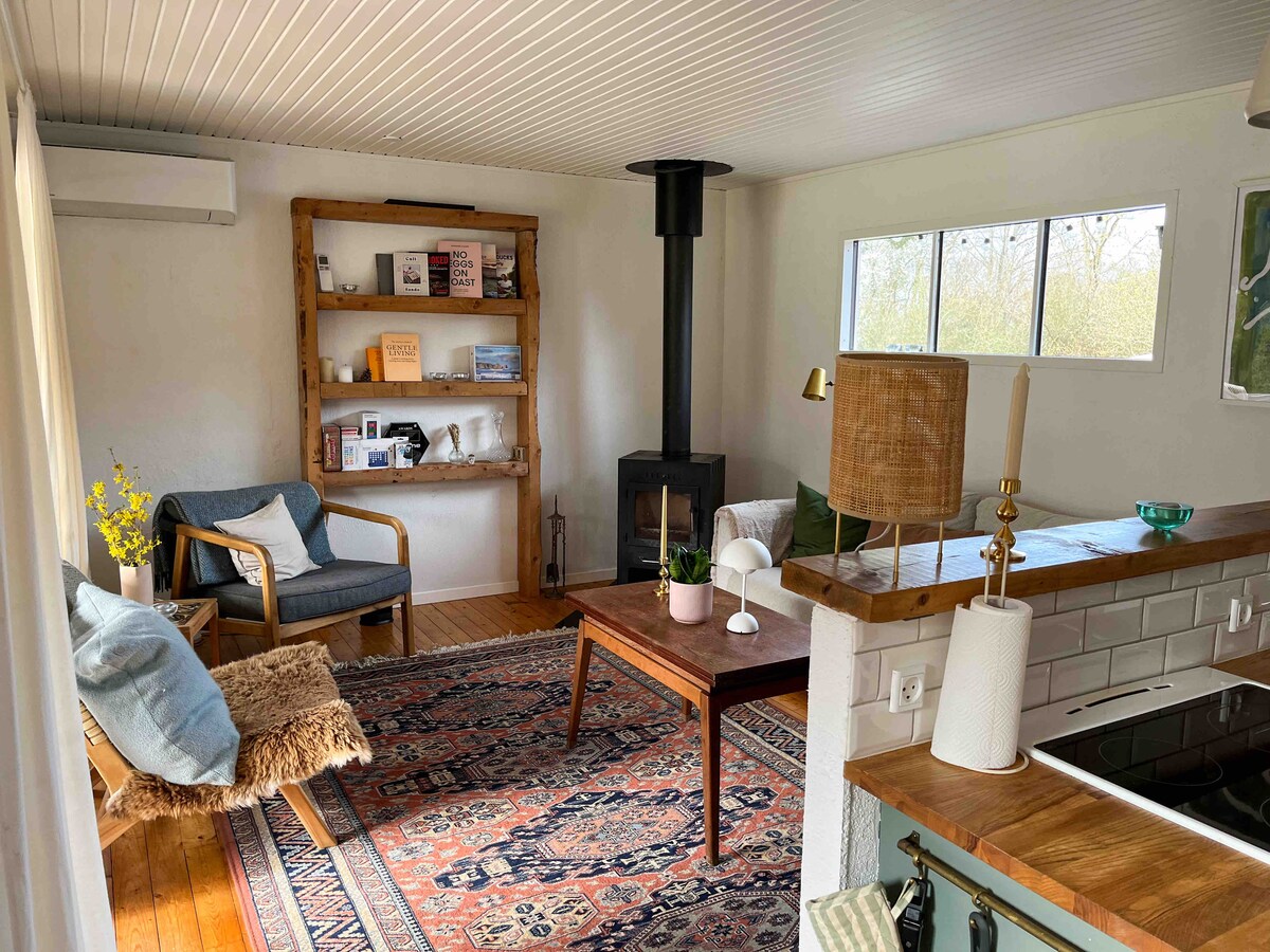 A bright living area features a cozy seating arrangement, including an armchair and a stylish rug. A wood-burning stove is positioned next to a wooden bookshelf filled with magazines. A spacious coffee table occupies the center, with natural light streaming in through the windows.