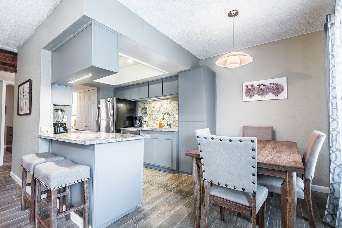 A modern kitchen and dining area are visible, featuring a granite countertop with bar seating for three. The dining table accommodates four. Neutral tones are complemented by decorative wall art and soft lighting, enhancing the space's overall functionality.