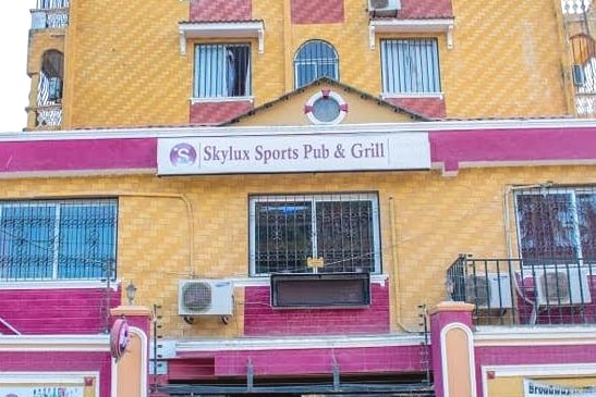 A vibrant building exterior displays the sign for Skylux Sports Pub & Grill. Large windows are visible, allowing natural light to enter. The facade features a mix of yellow and pink tones, contributing to its lively appearance.