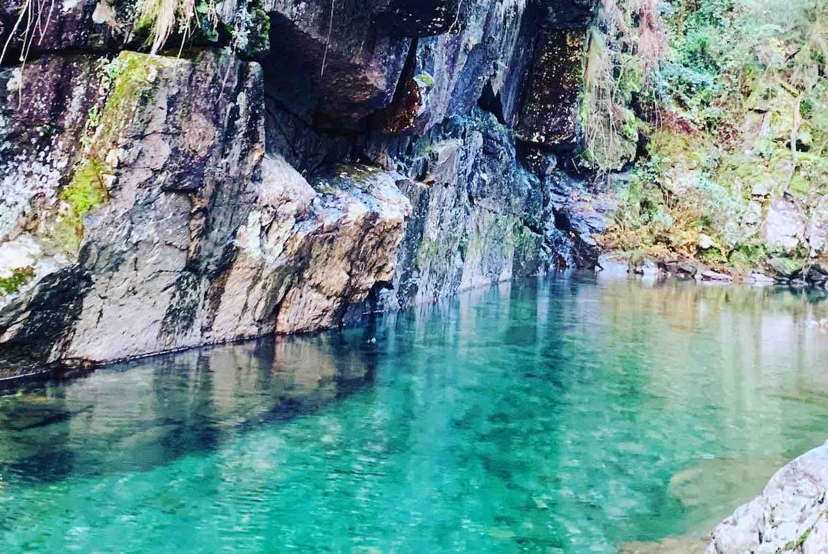 A serene scene of clear, turquoise water reflecting the surrounding rocks and greenery. The water appears calm, bordered by moss-covered stones and dense foliage, creating a peaceful atmosphere ideal for relaxation and nature appreciation.