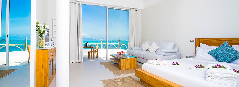 A spacious suite features floor-to-ceiling glass doors that provide stunning ocean views. The living area includes a plush leather sofa, a wooden coffee table adorned with flowers, and a king-sized bed draped in fresh linens. Natural light fills the room, creating a bright atmosphere.
