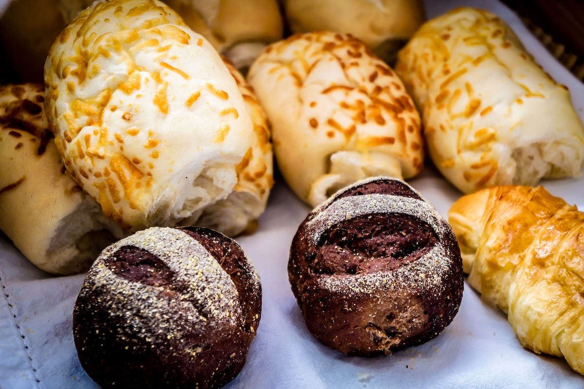 An assortment of freshly baked bread rolls is presented on a white cloth. Various types are visible, including soft rolls topped with cheese and darker rolls with a grainy texture. A golden croissant is also included, showcasing a variety of options for breakfast.