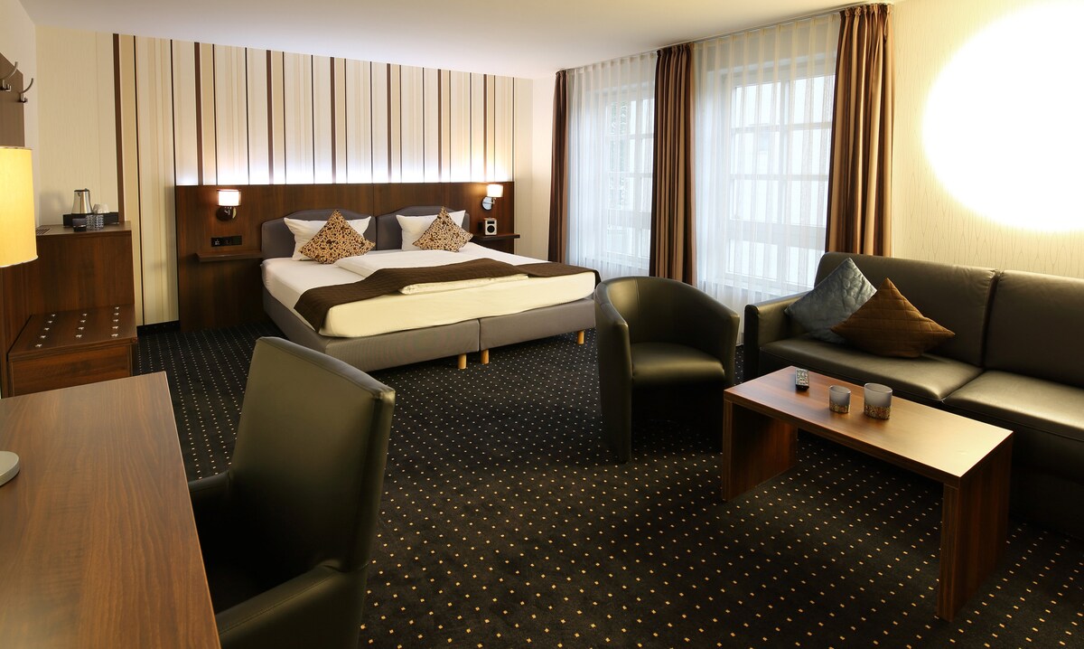 A spacious room featuring a comfortable bed with decorative pillows. A seating area is present with two chairs and a small table. Natural light enters through large windows, and the decor includes vertical stripes and warm tones, contributing to an inviting atmosphere.