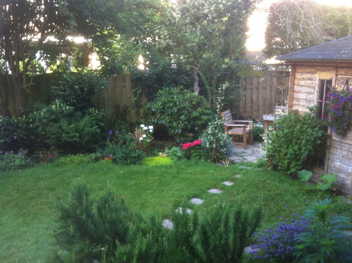 A spacious garden features well-manicured grass, bordered by various plants and flowers. Stone stepping stones lead through the greenery towards a wooden shed, while a seating area is nestled among the lush foliage, providing a serene outdoor space.