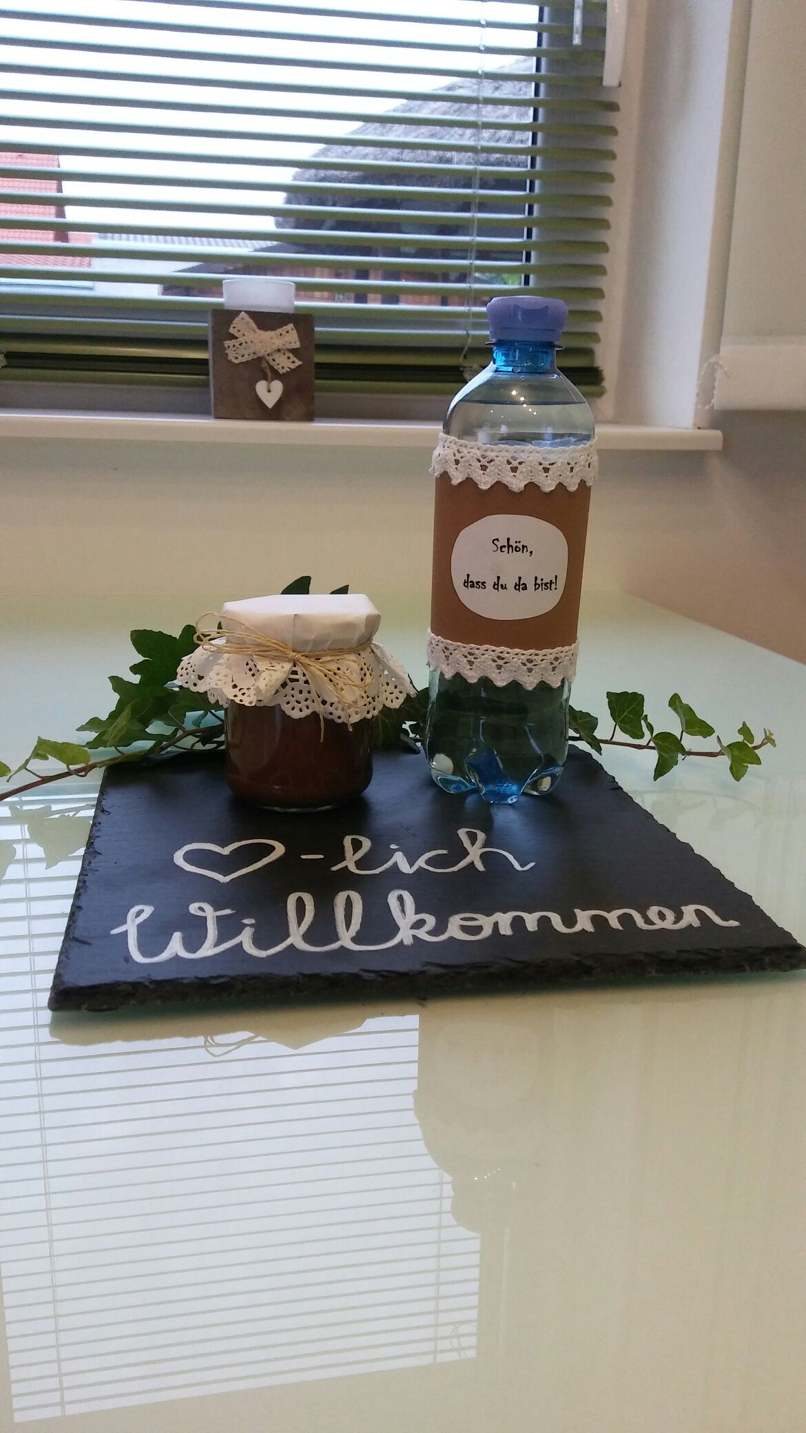 A welcoming display is arranged on a slate platter featuring a jar of homemade jam and a bottle of water, both adorned with decorative labels. A small potted plant adds a touch of greenery, while a handwritten note conveys a message of hospitality.