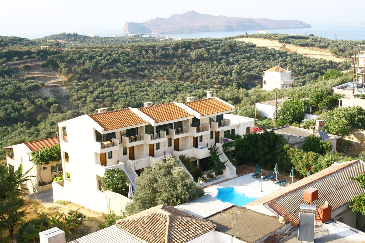 The hotel complex is viewed from above, showcasing multiple two-story buildings surrounded by lush greenery. A swimming pool is centrally located, with loungers nearby. The background features rolling hills and the coastline, with the sea visible in the distance.
