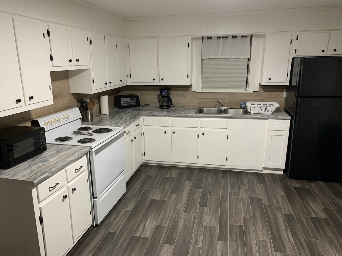 A spacious kitchen showcases white cabinetry and a large countertop with a subtle gray pattern. Essential appliances are provided, including a stove, oven, microwave, and refrigerator. Natural light filters through the window, illuminating the tile floor that adds warmth to the room.