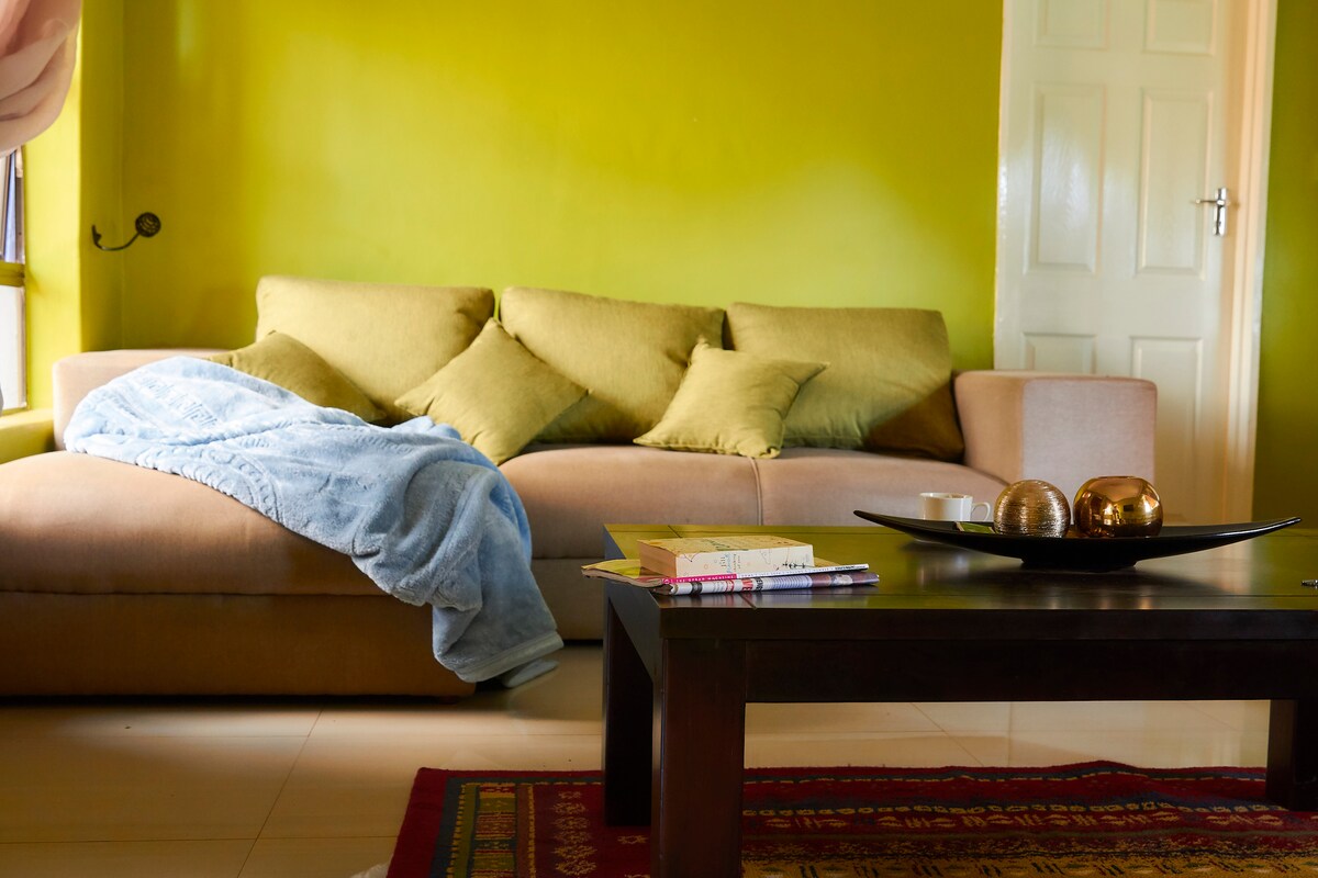 A comfortable sectional sofa in green and beige tones is placed against a bright green wall. The sofa is adorned with several cushions. A coffee table with decorative items rests in front of the sofa, while natural light illuminates the tranquil living space.