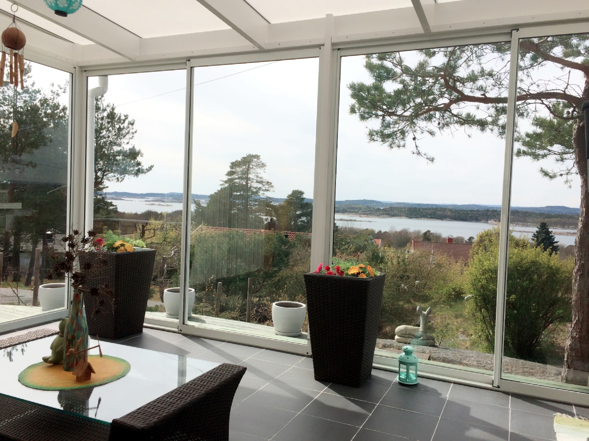 A sunlit sunroom features large glass windows that frame a panoramic view of the surrounding landscape, including water and distant trees. Two planters with colorful flowers add a touch of greenery to the space. A tranquil atmosphere is created by the open and airy design.