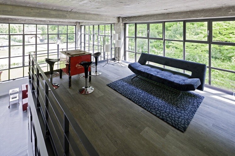 An open gallery space features large windows allowing natural light to illuminate the area, complemented by views of greenery outside. A modern sofa is situated on a textured rug, alongside a round table and slender stools, providing a minimalist ambiance.