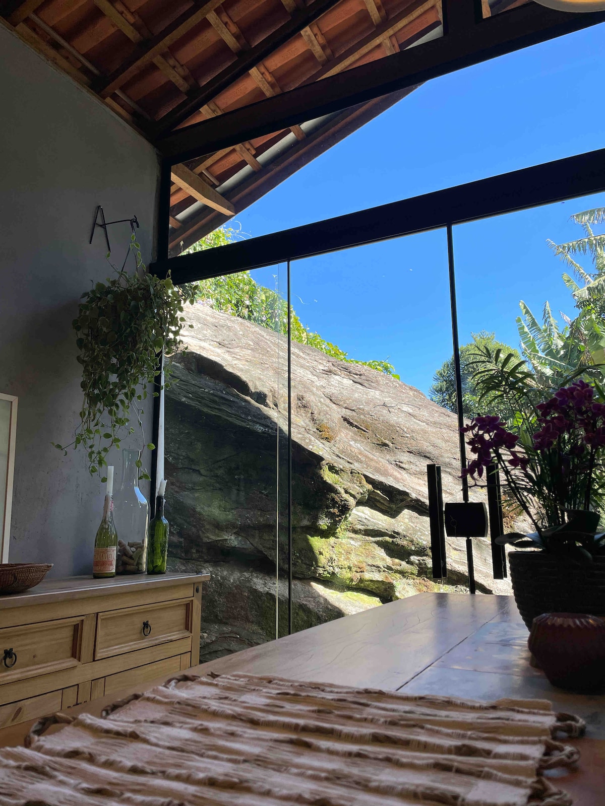 A spacious interior is highlighted by large glass doors that offer unobstructed views of a natural rock formation and surrounding greenery. Light enters the room, enhancing the simplicity of a wooden dining table adorned with a patterned table runner and potted plants.