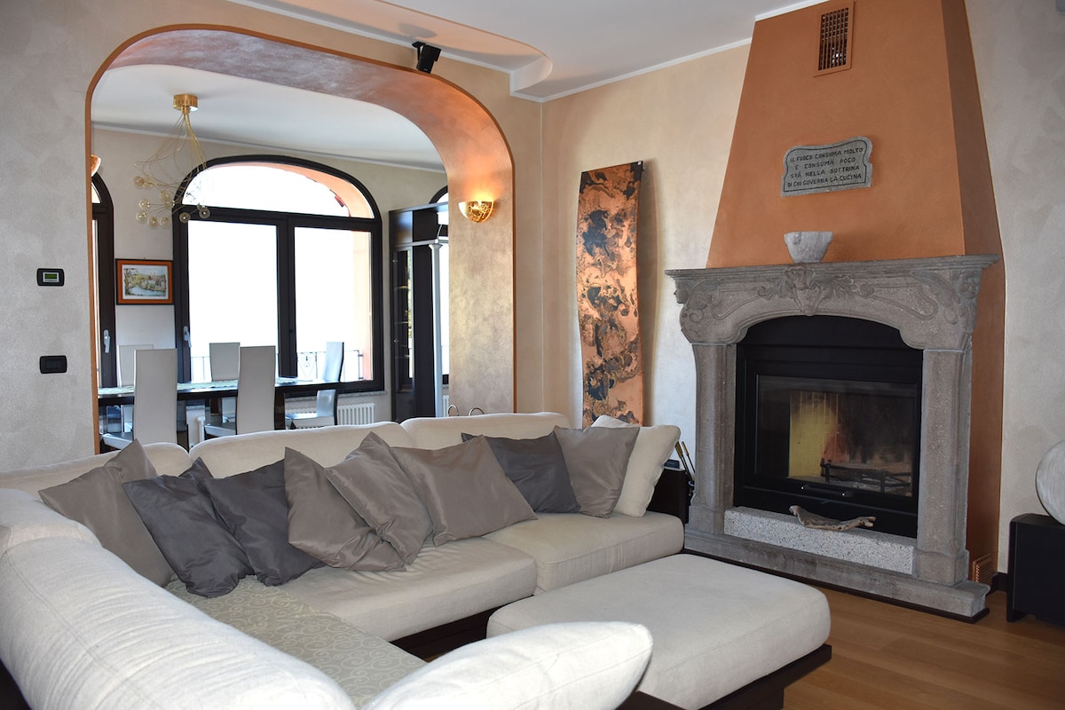 The spacious living area features a large, U-shaped sofa adorned with gray pillows. A stone fireplace is situated under an arched alcove, complemented by a piece of wall art nearby. Natural light floods the room through large windows, enhancing the inviting atmosphere.