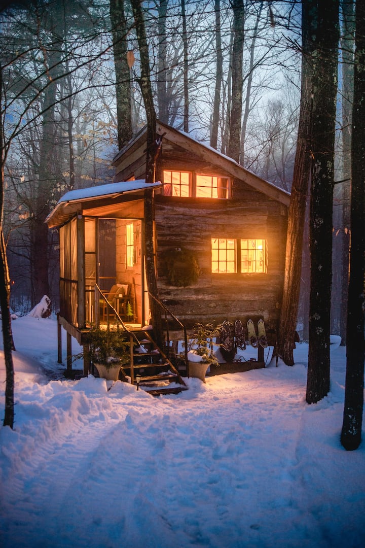A tiny house rental in New Hampshire is seen lit up with warm lights from within on a snowy day