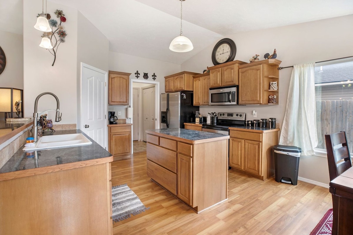 A spacious kitchen features wooden cabinetry and dark countertops, complemented by a central island. Modern appliances, including a refrigerator and stove, are visible. Natural light enters through a nearby window, highlighting the open layout and inviting atmosphere, with a door leading to additional living areas.