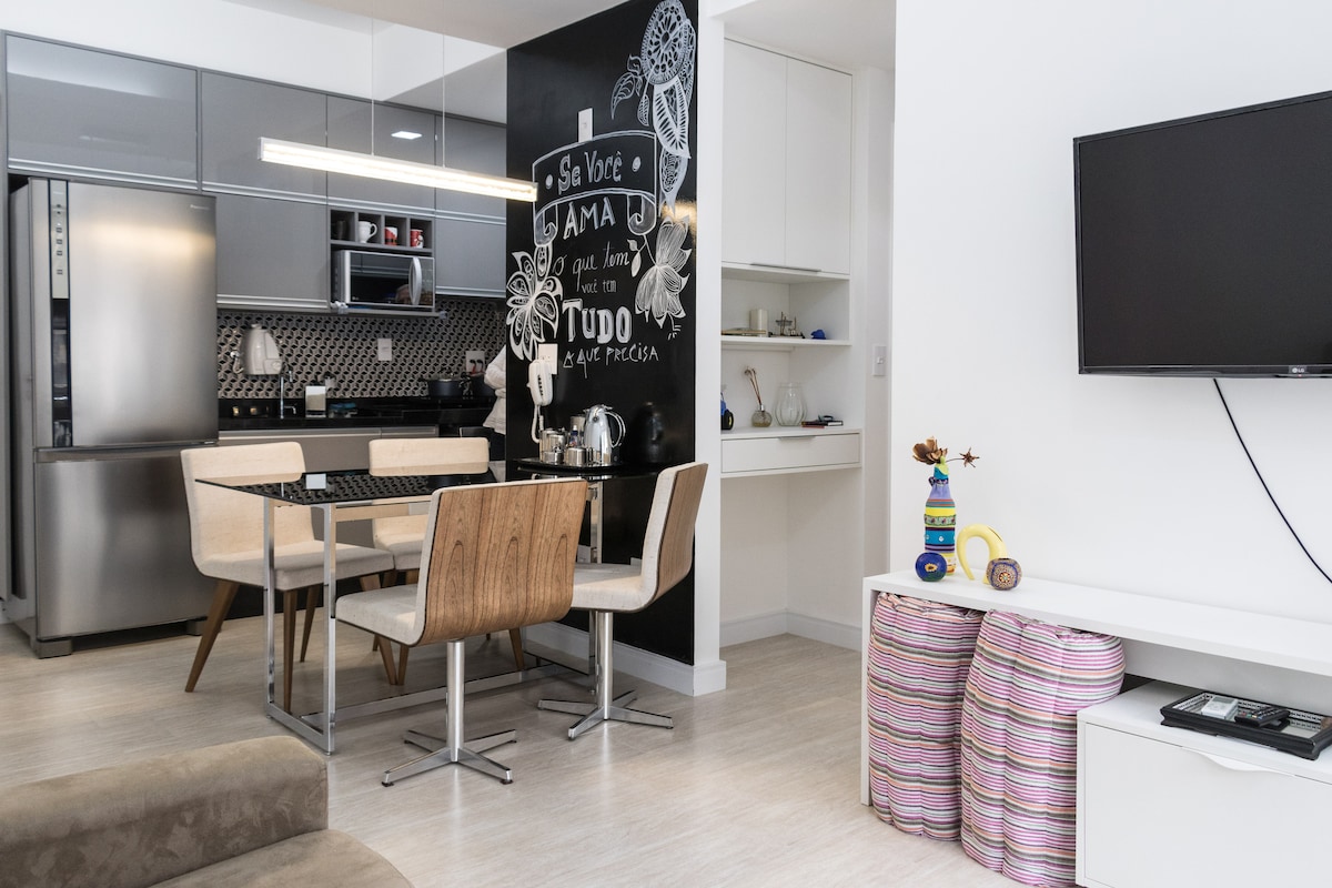 A modern kitchen and dining area is visible, featuring a large refrigerator and sleek cabinetry. The dining table is surrounded by four wooden chairs. A chalkboard wall adds a creative touch. A flat-screen TV is mounted on the wall, complementing the minimalist decor.