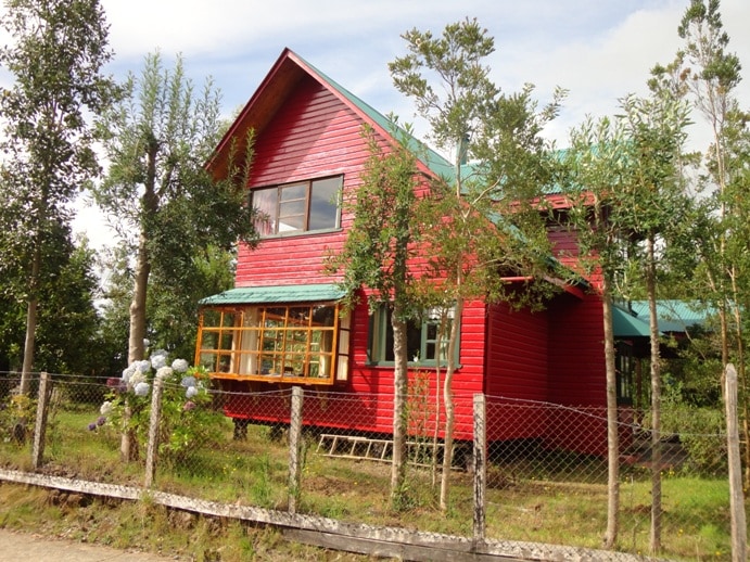 A red wooden cabin is surrounded by greenery, showcasing large windows that allow natural light to fill the interior. The structure has a gabled roof and is complemented by a row of bright flowers nearby. A wooden fence lines the front of the property.