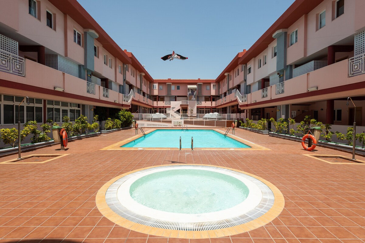 A central outdoor pool area is surrounded by two-story accommodations. The pool is complemented by a circular hot tub and inviting lounge spaces. Sunlight reflects off the water, and colorful planters add greenery to the tiled deck. Lifebuoys are positioned at various points for safety.