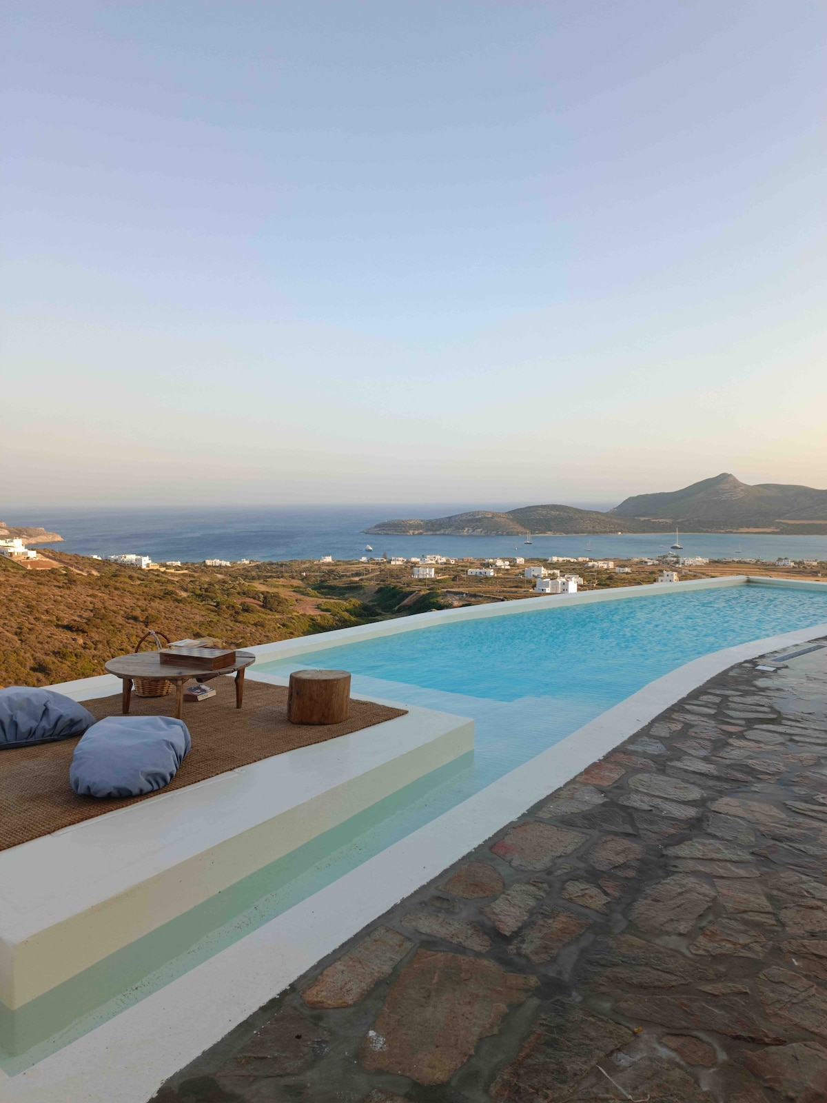 An infinity pool is seen extending towards the horizon, with the tranquil sea and distant islands in view. Comfortable seating arrangements are positioned nearby, set against a backdrop of natural landscapes and the soft evening light.