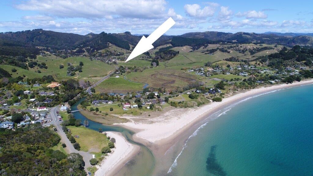 An aerial view captures the serene landscape of Kuaotunu, showcasing the winding river leading to the shore. The sandy beach meets the turquoise water, with rolling hills in the background and scattered homes dotting the coastline, highlighting the area's natural beauty.
