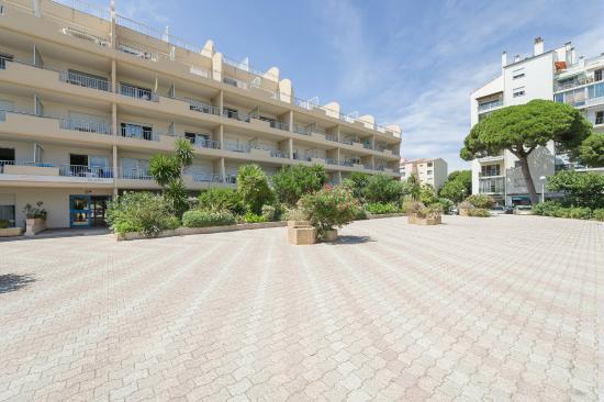 The image displays a spacious courtyard in front of a multi-story residential building. Lush greenery and potted plants create a relaxed atmosphere, while the building's multiple balconies add architectural interest. The well-maintained paving stones provide a clear pathway leading to the entrance.