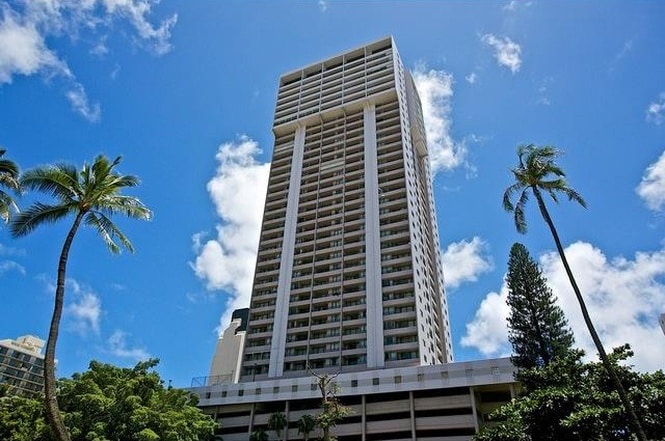 A tall condominium building rises against a bright blue sky, featuring multiple floors and a modern architectural design. Lush palm trees and vibrant green foliage surround the base, creating an inviting coastal atmosphere.