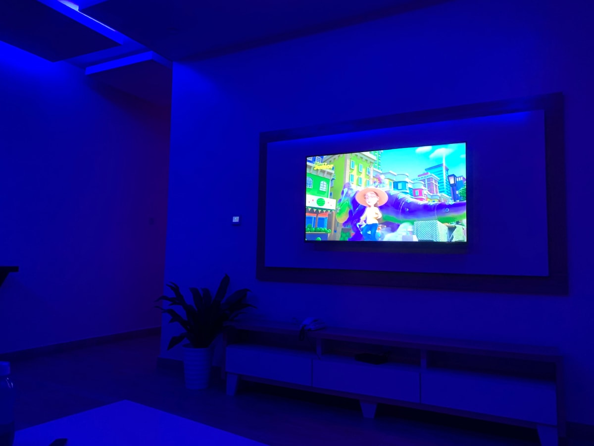 A lounge area features a large wall-mounted television displaying vibrant animated content. Ambient blue lighting bathes the room, enhancing the modern decor. A potted plant adds a touch of greenery, and a sleek media console sits below the TV, providing storage and style.