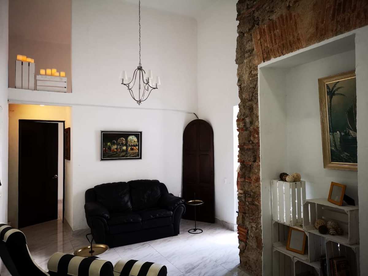 A sunlit living area features a black leather couch and contrasting striped chairs. A chandelier hangs from the high ceiling, with a partially visible staircase leading to a loft above. Exposed brick accents and a piece of artwork contribute to the modern yet historic ambiance.