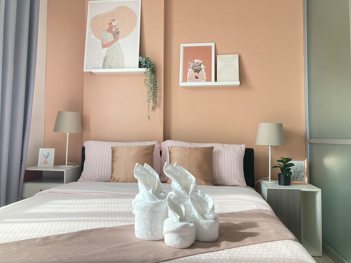 A cozy sleeping area features a spacious bed with patterned bedding and accent pillows. Decorative wall art is displayed above the bed, complemented by a soft color palette. Two table lamps provide gentle lighting, and plush slippers are arranged neatly on the bed.