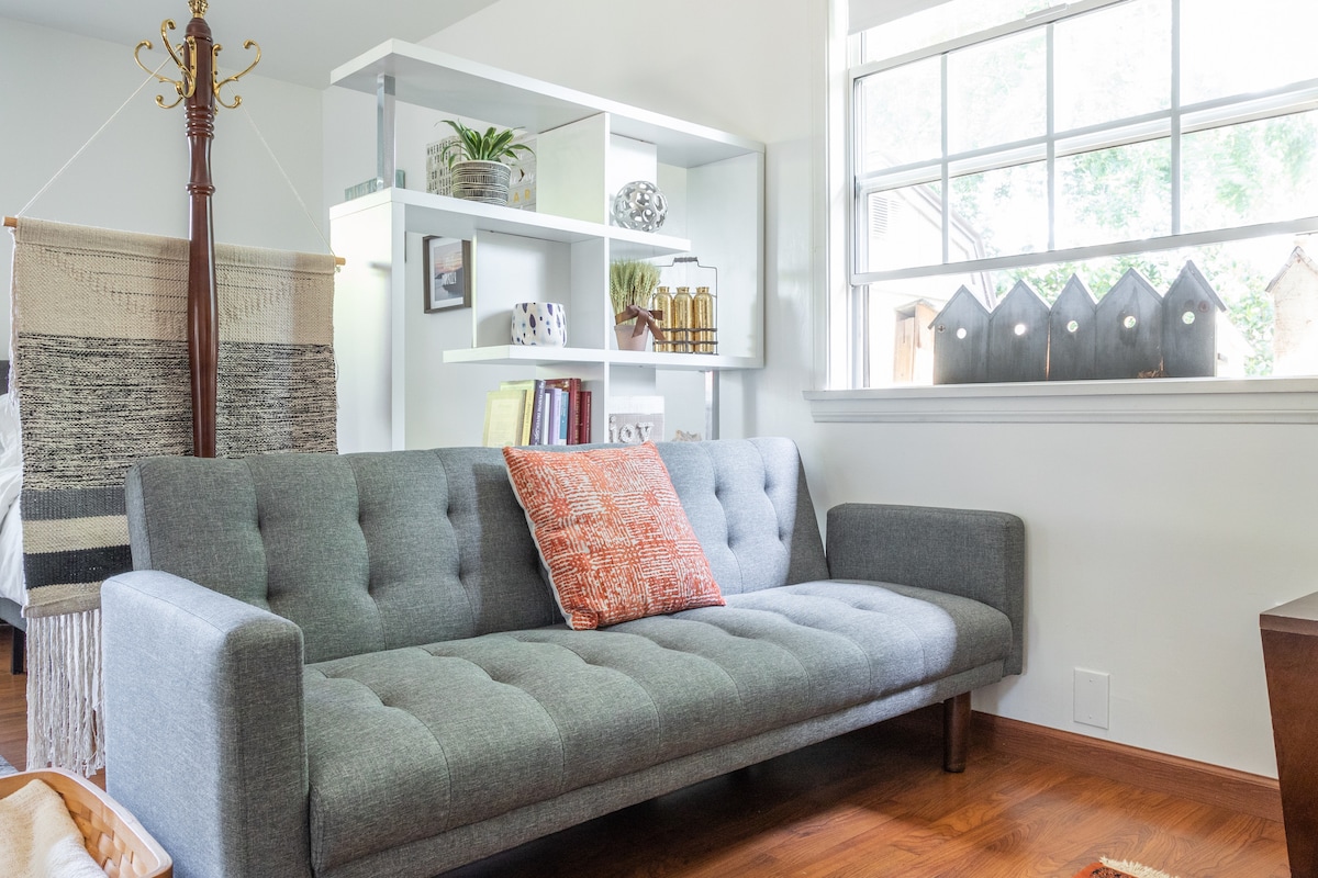 A cozy seating area is featured with a modern gray futon, accented by a rust-colored decorative pillow. Natural light pours through a large window, illuminating a shelf filled with potted plants and decorative items. A wooden accent table complements the space.
