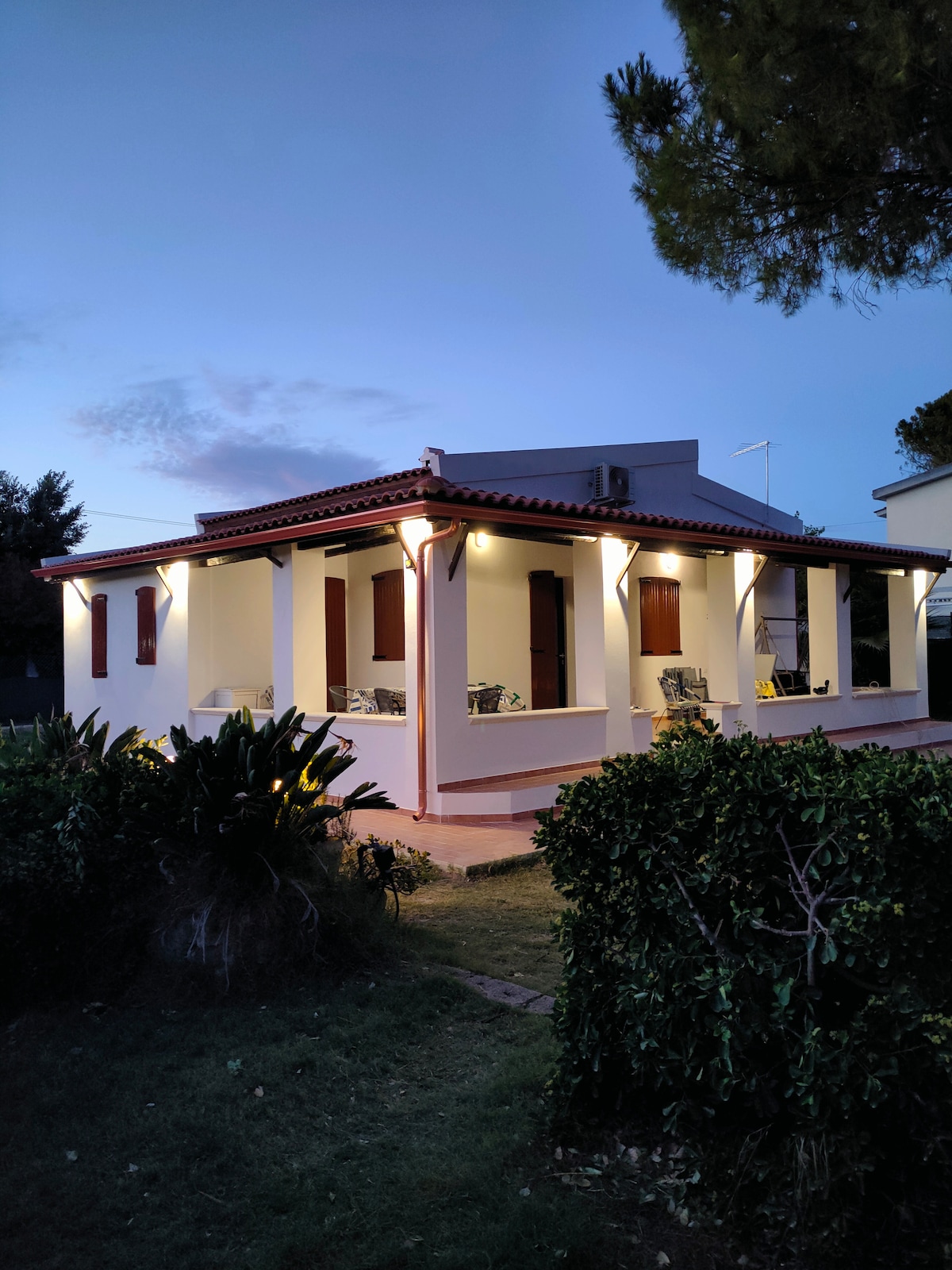The exterior of a charming house is illuminated at twilight, featuring a covered veranda and large windows. Lush greenery surrounds the property, enhancing the inviting atmosphere. The architectural style combines traditional elements with modern touches, providing an ideal setting for relaxation.