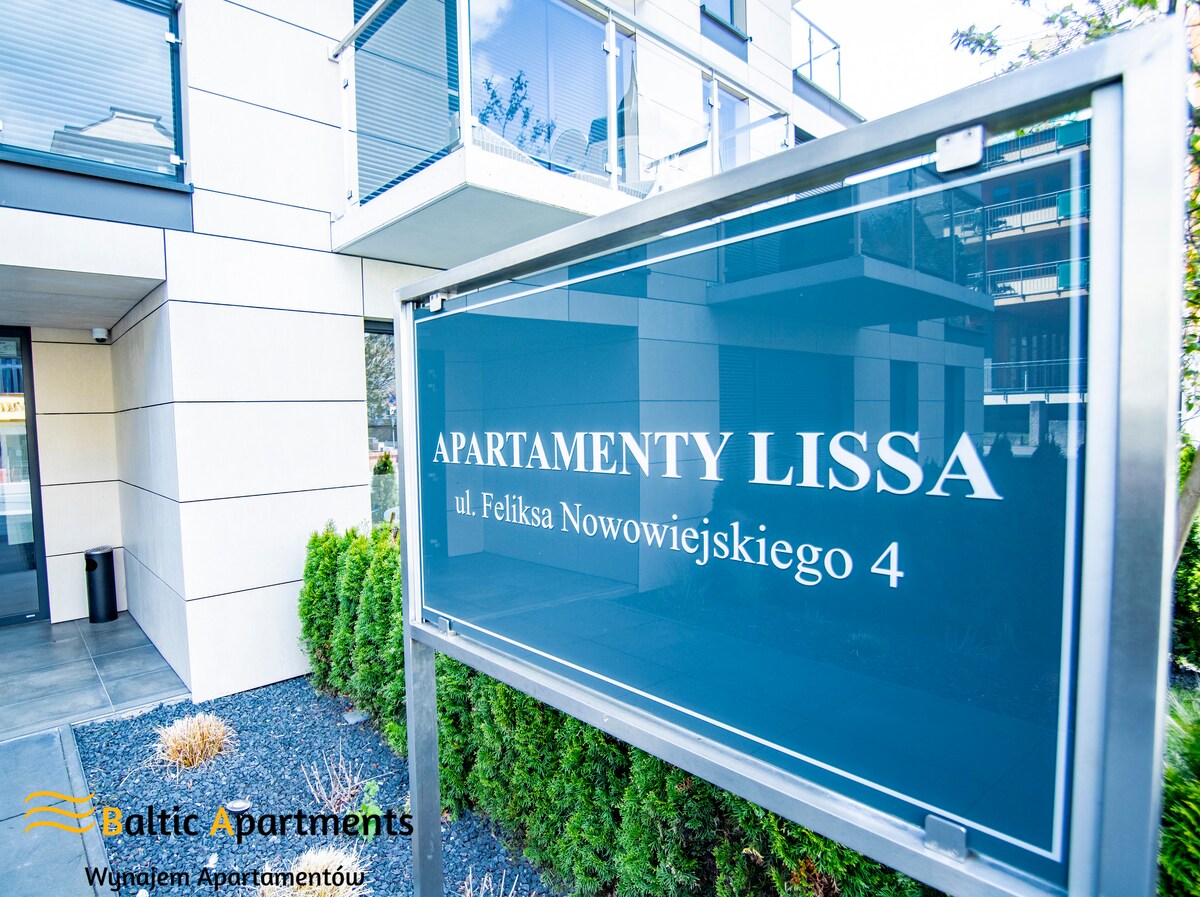 A modern building features a prominent entrance sign displaying 'Apartamenty Lissa.' The exterior is characterized by clean lines and a contemporary design, surrounded by neatly landscaped greenery that enhances the welcoming appearance.