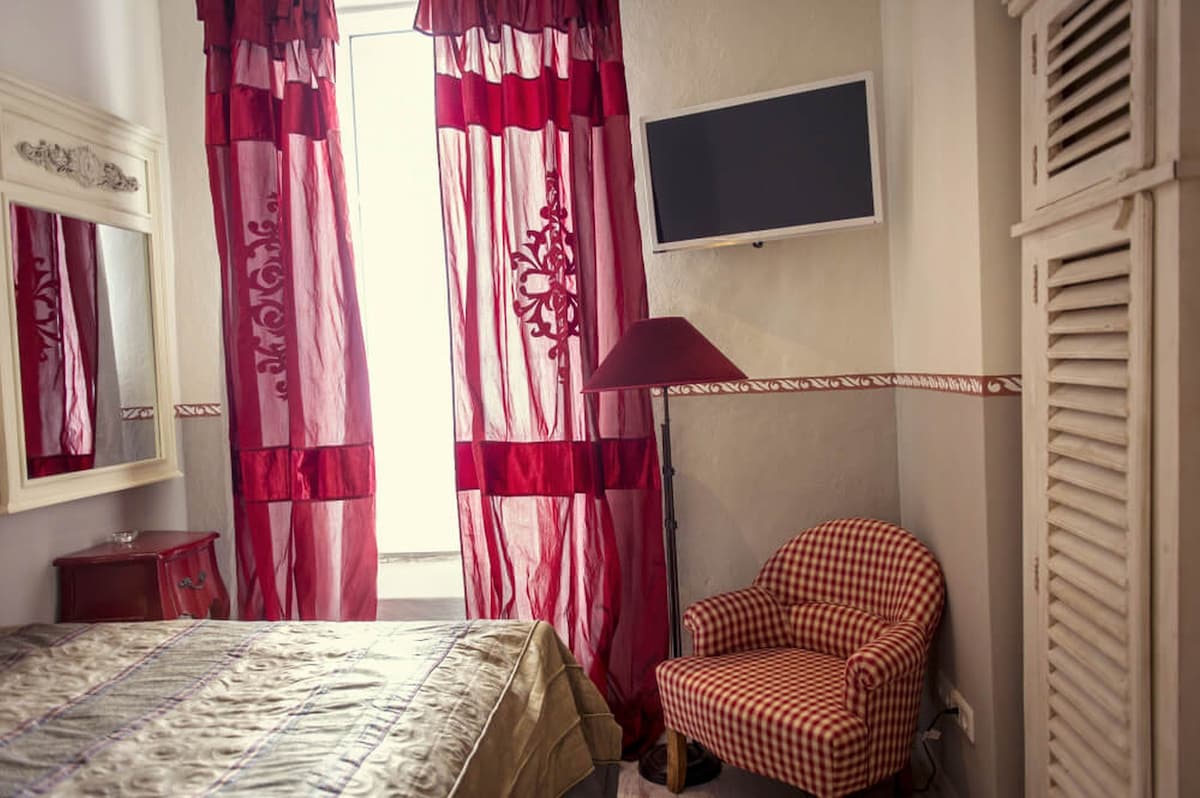 A room features a spacious bed with a patterned cover, complemented by bright red striped curtains that frame a window. A compact television is mounted on the wall, and a small checked armchair is positioned nearby, adding to the cozy ambiance.