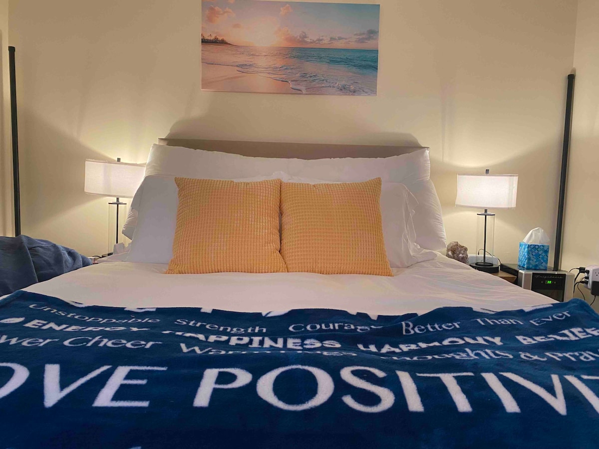 A serene bedroom is highlighted by a spacious bed covered with a blue blanket featuring inspirational words. Two yellow pillows are positioned at the head of the bed, alongside modern bedside lamps. A tranquil beach scene is displayed on the wall above, contributing to a calming atmosphere.