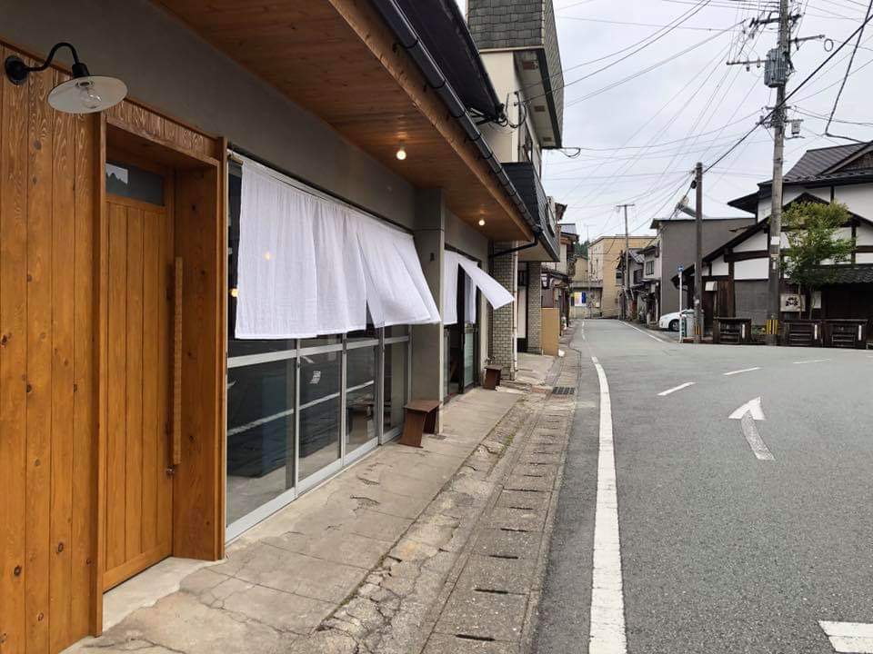 A traditional building features a wooden entrance and large windows adorned with white curtains. The exterior blends with the surrounding street, showcasing a mix of modern and historic architecture. A sidewalk runs parallel, with scattered street signs and power lines visible.
