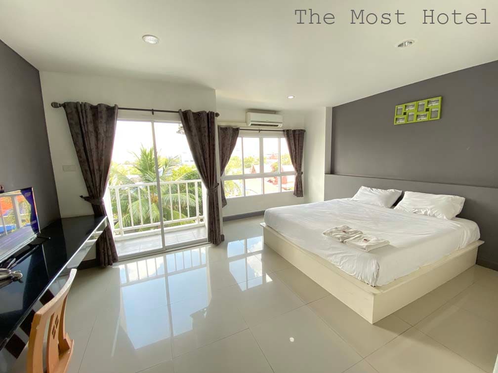 A bright and spacious room features a large bed with fresh linens, complemented by a seating area with a desk. Natural light fills the space through floor-to-ceiling windows, which offer views of greenery. Dark curtains frame the windows, while air conditioning and a TV are also present.
