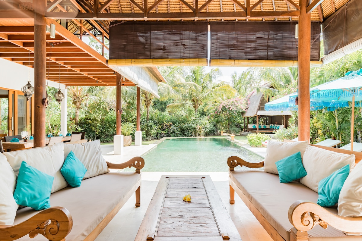 A spacious lounge area features wooden seating with plush cushions in shades of turquoise. Natural light floods in, highlighting views of the lush garden and a glistening pool in the background, framed by tropical plants and serene lounge chairs.