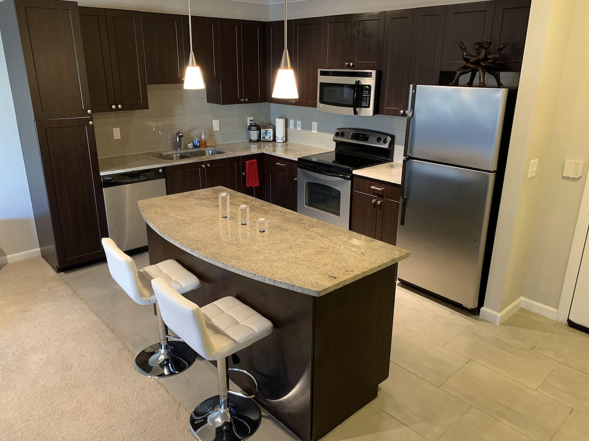 A modern kitchen is presented with dark wood cabinetry and a light-colored countertop. Bar stools are positioned at a curved island, while stainless steel appliances, including a refrigerator and oven, are neatly arranged. Pendant lighting adds soft illumination to the work area.