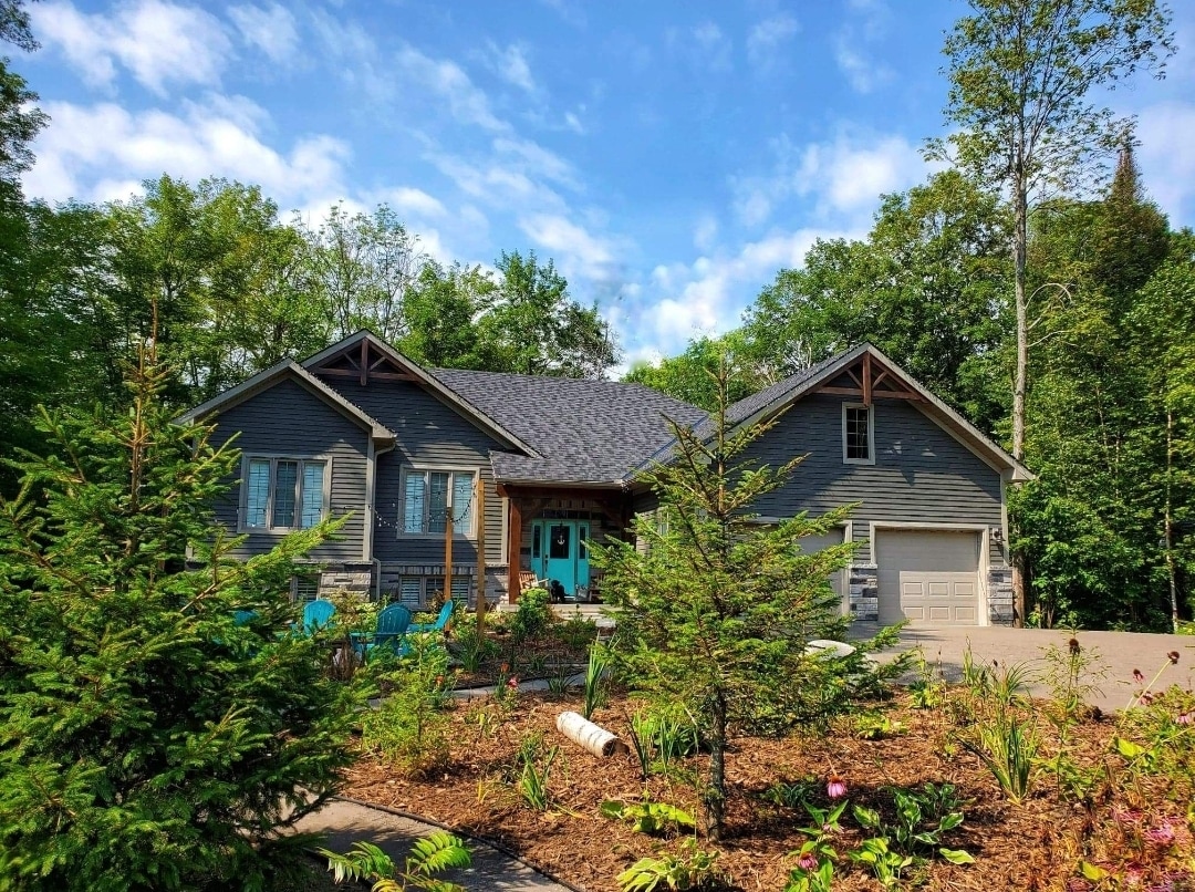 The exterior of a spacious bungalow is surrounded by lush greenery and colorful landscaping. A well-maintained driveway leads to the garage, and cozy outdoor seating is placed near the entrance, inviting relaxation among natural elements.