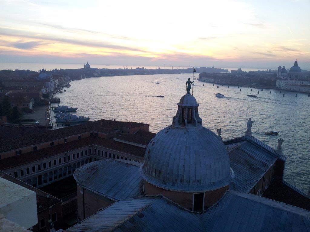 A panoramic view of a tranquil Venetian waterway at dusk, highlighted by the soft glow of the setting sun. The silhouette of historic buildings and distant islands is visible against the colorful sky, creating a picturesque backdrop for the serene waters below.