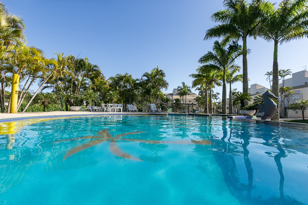 A clear swimming pool is surrounded by lush palm trees and vibrant greenery. Comfortable lounge chairs are positioned near the poolside, providing a relaxing space for guests. Bright blue water reflects the clear sky, creating a refreshing atmosphere.