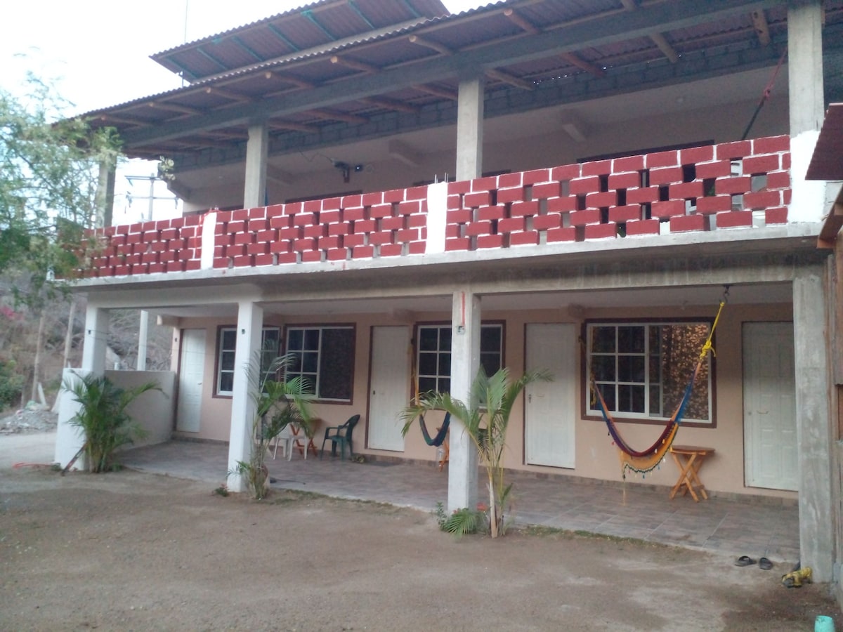 A multi-level building with an open-air design features a covered balcony supported by concrete pillars. The ground level showcases a series of doors leading to individual rooms, with a small table and chairs positioned on a tiled area. Lush greenery is present near the entrance.