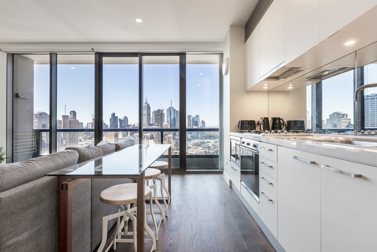 A modern kitchen and dining area is illuminated by natural light from large windows, offering stunning views of the city skyline. A sleek dining table with grey chairs is positioned next to a well-equipped kitchen featuring stainless steel appliances and a contemporary layout.