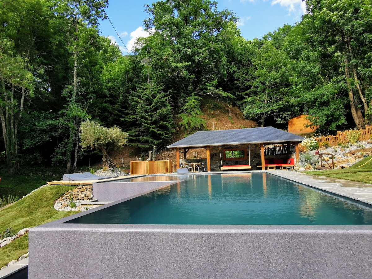 A modern infinity pool is surrounded by lush greenery, seamlessly integrating with the natural landscape. A wooden deck and a covered lounge area with a black roof offer relaxation options. Mature trees provide shade, enhancing the outdoor ambiance.